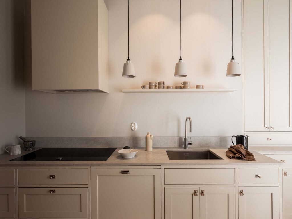 Stiligt - Minimalistisk beige Shakerstil kök med spishäll, diskbänk och bänkskiva. Tre taklampor hänger ovanför; hyllor rymmer fat och koppar. En vattenkokare och en handduk vilar på bänken.