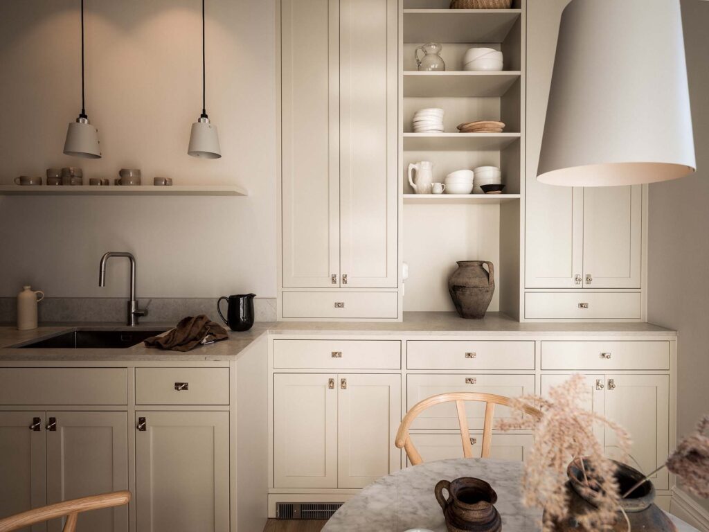 Stiligt - Ett modernt kök med krämskåp, en stiligt bänkskiva i marmor, öppna hyllor med porslin, ett handfat, en svart vattenkokare och pendelbelysning över ett runt bord. Färgen för ditt platsbyggda kök
