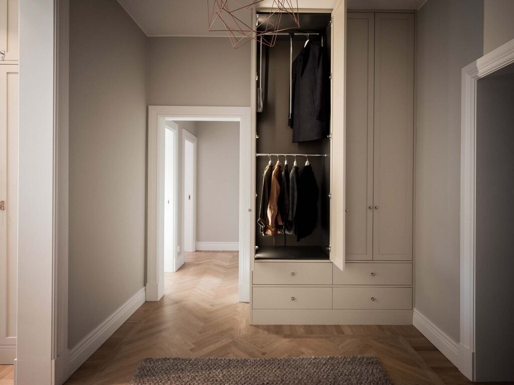 Stiligt - En minimalistisk garderob med öppna dörrar visar flera jackor och skjortor som hänger inuti, lådor under, och en geometrisk armatur ovanför i en neutral tonad hall.