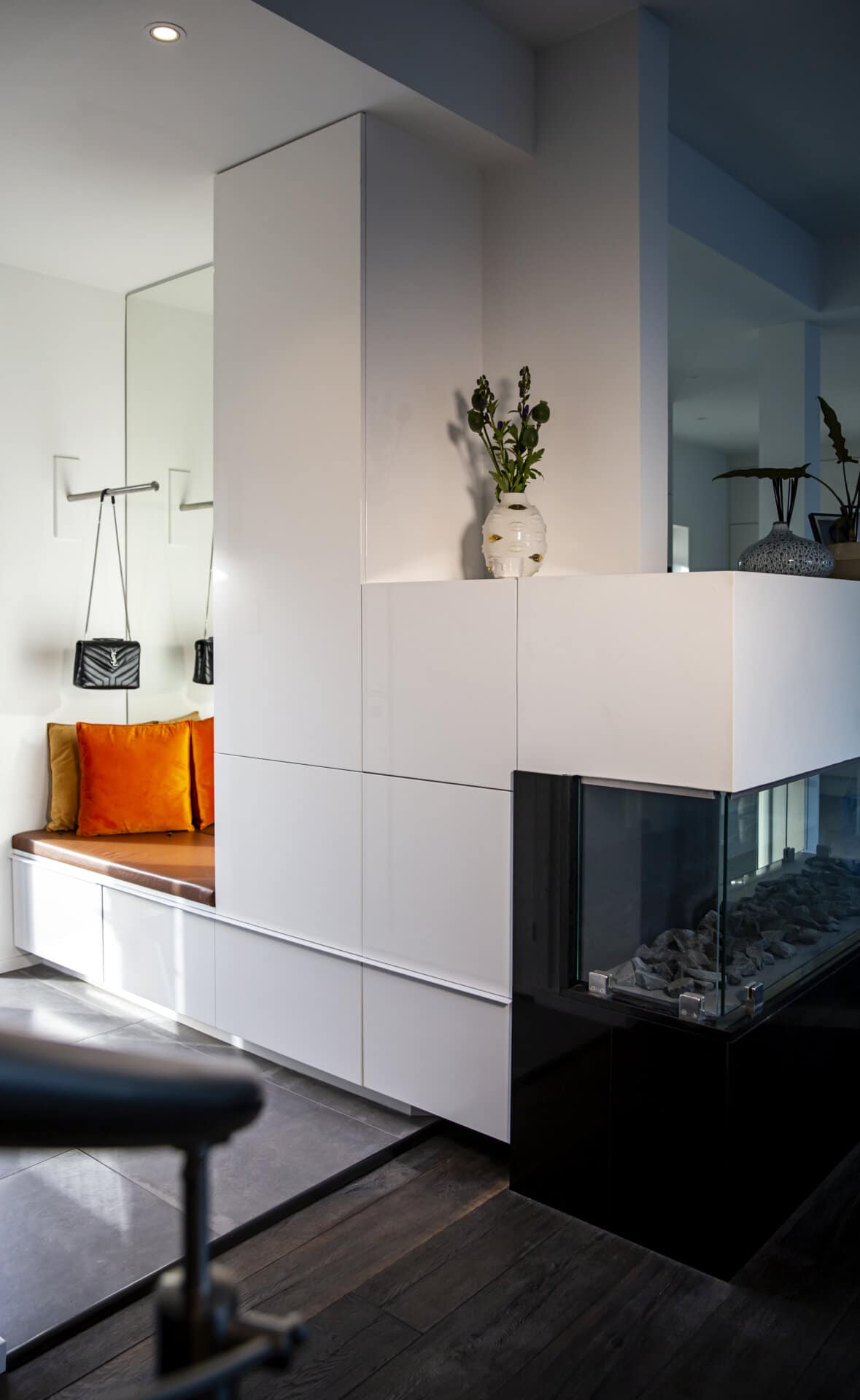 Stiligt - Modern inredning med vitt inbyggt skåp, inglasad öppen spis, orange-kuddad bänk och väggkrokar som håller påsar, med växter på skåpet.