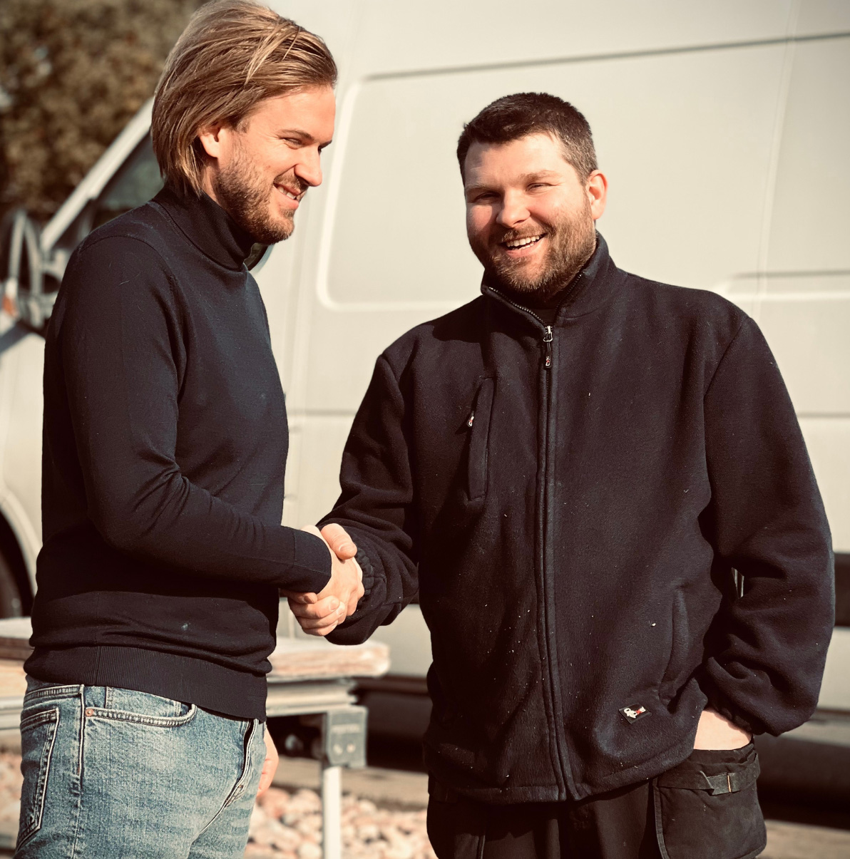 Stiligt - Två män står utomhus framför en vit skåpbil, skakar hand och ler mot varandra.