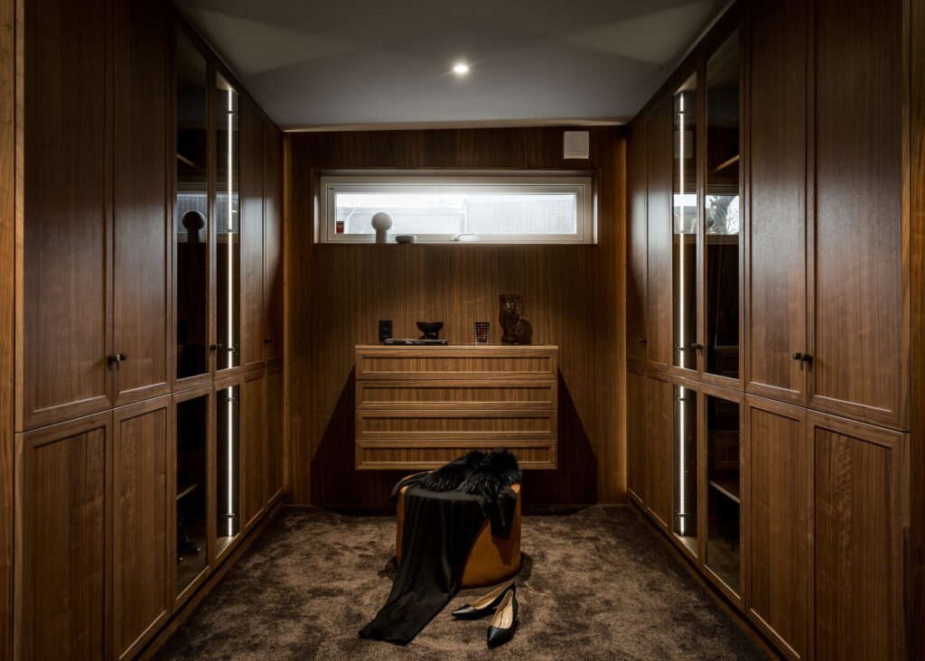Stiligt - En klassisk walk in closet med träskåp, en central byrå, ett fönster ovanför och en brun ottoman draperad med kläder sitter elegant på det heltäckningsbelagda golvet bredvid ett par skor.
