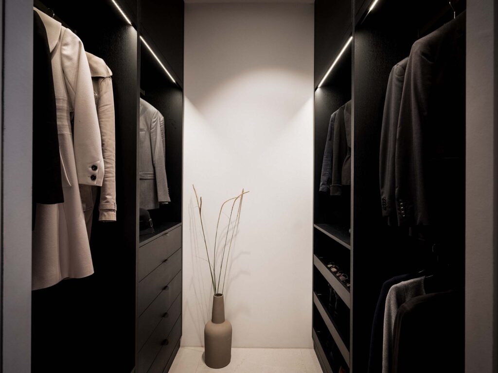 Stiligt - En modern liten walk in closet med mörka hyllor, hängande jackor och skjortor, lådor och en dekorativ vas med högt torkat gräs i mitten.