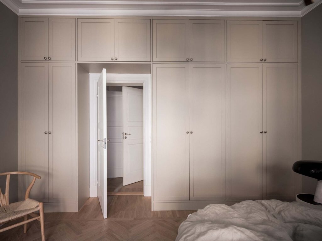 Stiligt - Ett sovrum med en garderobsvägg i sovrum med inbyggda beige garderober som ramar in en dörröppning, en delvis öppen vit dörr, en trästol och en obäddad säng med vita sängkläder i förgrunden.