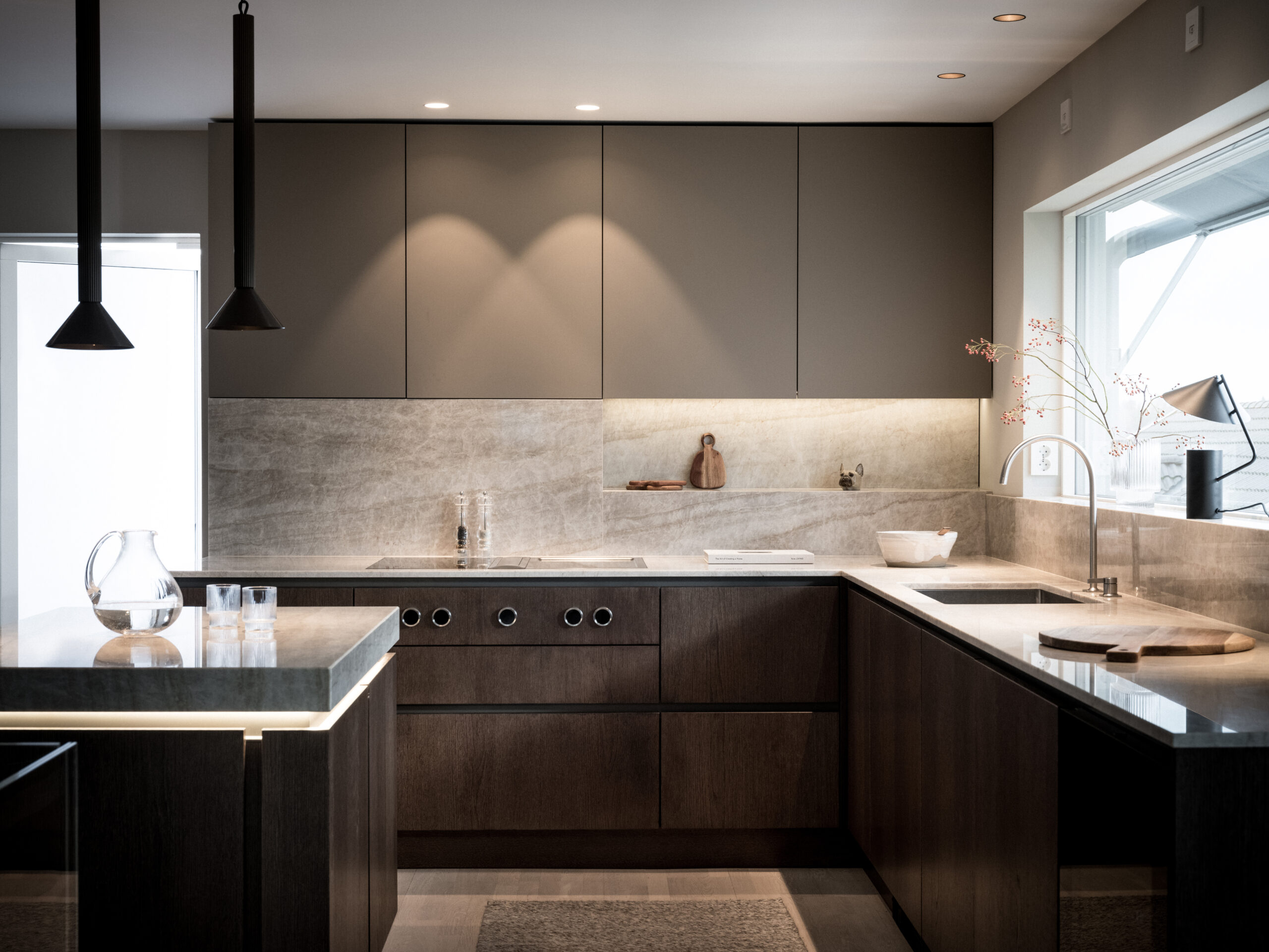 Stiligt - Ett modernt kök med stiligt mörka träskåp, bänkskivor i marmor och snygga svarta taklampor. Den har en minimalistisk design som accentueras av en vas och skärbräda på bänken.