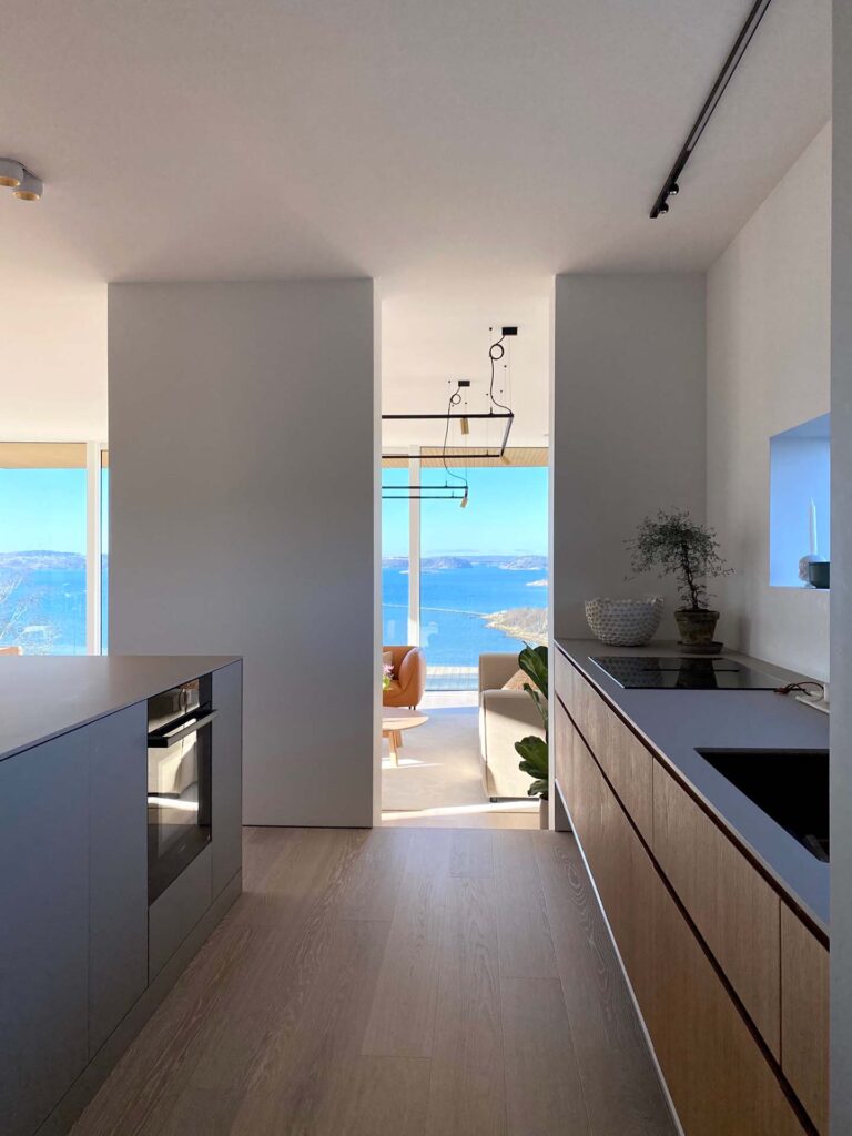 Stiligt - Modernt kök med trä och vita skåp, inspirerat av Villa Fernström och Husdrömmar, leder till ett ljust vardagsrum med stora fönsterpartier och utsikt över havet och kusten utanför.