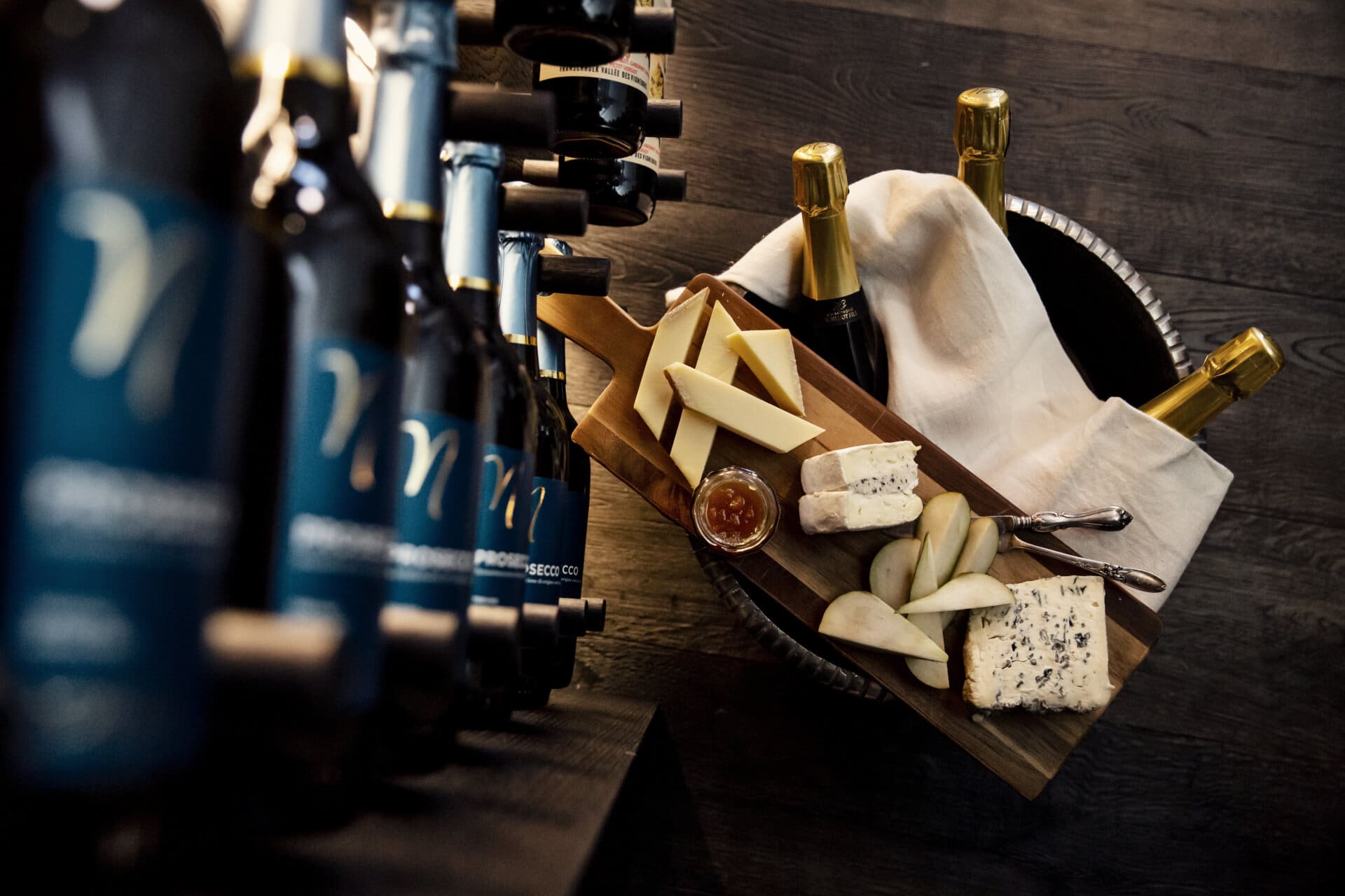 Stiligt - En träskiva med diverse ostar, päronskivor och sylt står bredvid flaskor vin och champagne på ett mörkt träbord.