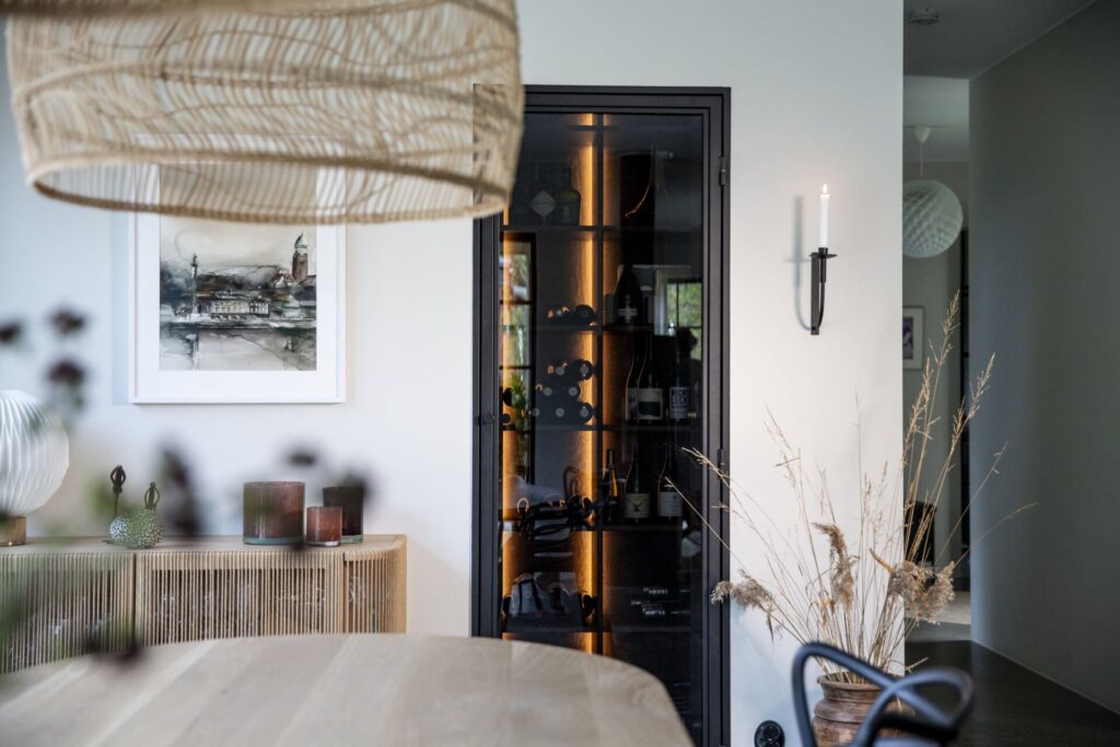 Stiligt - En modern matplats med ett träbord, flätad lampa, väggkonst, en dekorativ hylla, ett vinskåp med glasdörr och torkade växter i en vas.