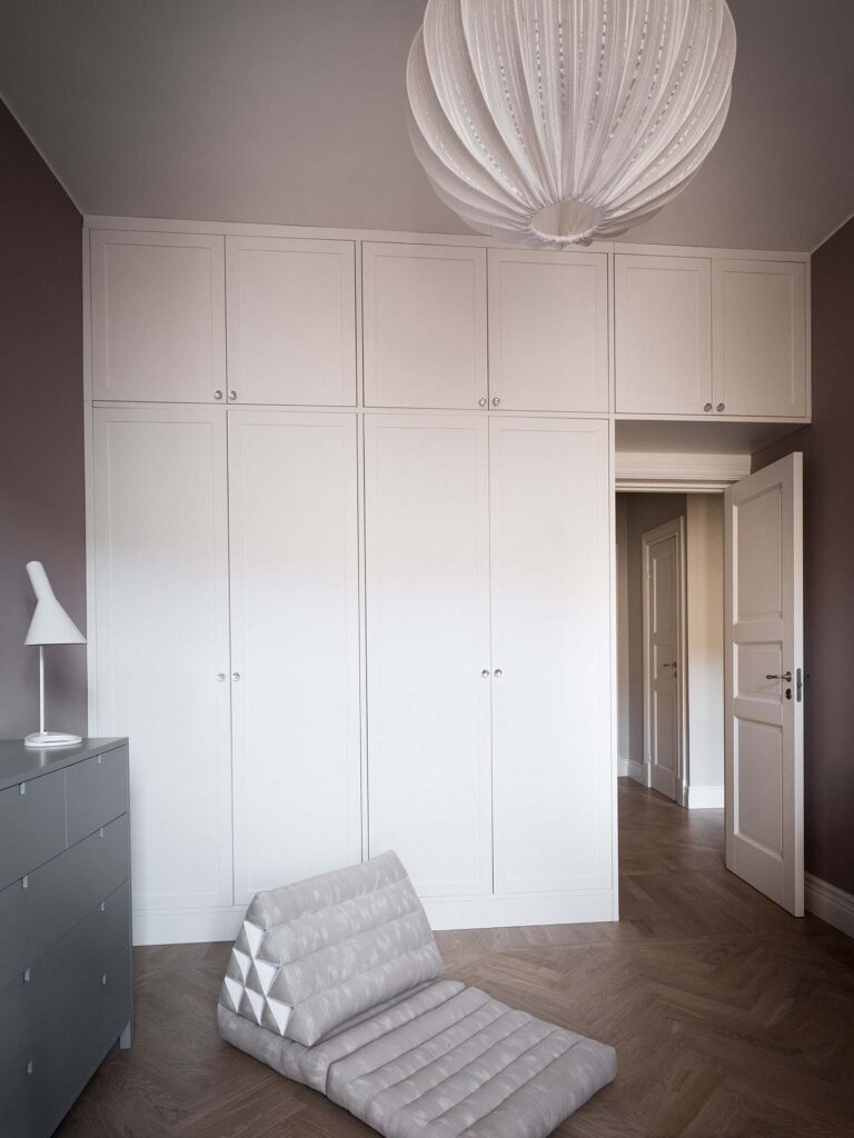 Stiligt - Minimalistiskt sovrum med en Vit Shakergarderob med städskåp, en grå byrå med en vit lampa, en beige hopfälld golvstol och en stor vit taklampa. Dörren till hallen är öppen.