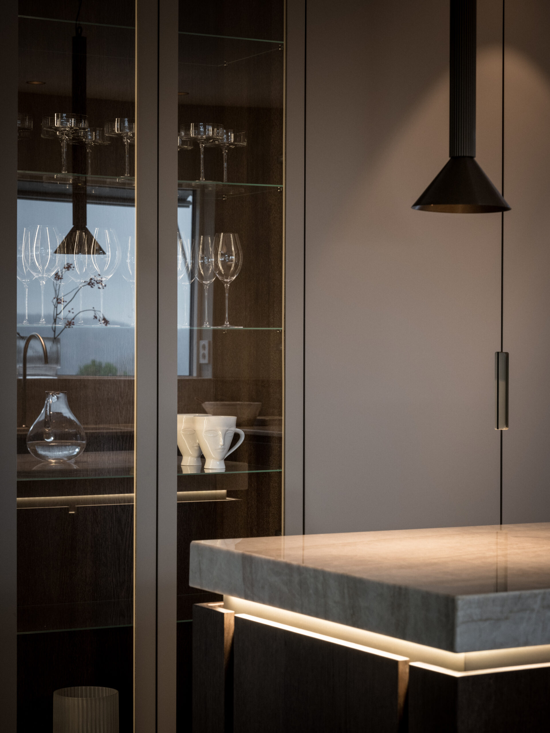 Stiligt - Modern köksinredning med ett stiligt vitrinskåp med vinglas och koppar, kompletterat med en snygg grå bänkskiva med underbelysning och en hängande svart taklampa.