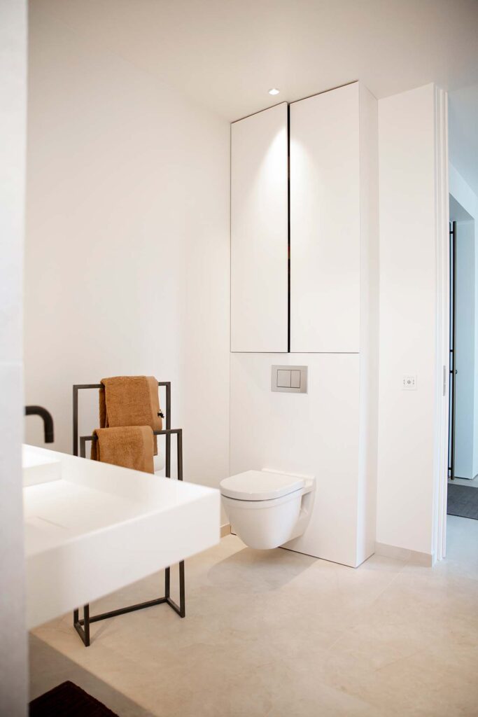 Stiligt - Villa Fernströms badrum Ett stiligt minimalistiskt badrum har vita väggar, en väggmonterad toalett, ett elegant vitt handfat och en handdukshållare i metall prydd med eleganta bruna handdukar.