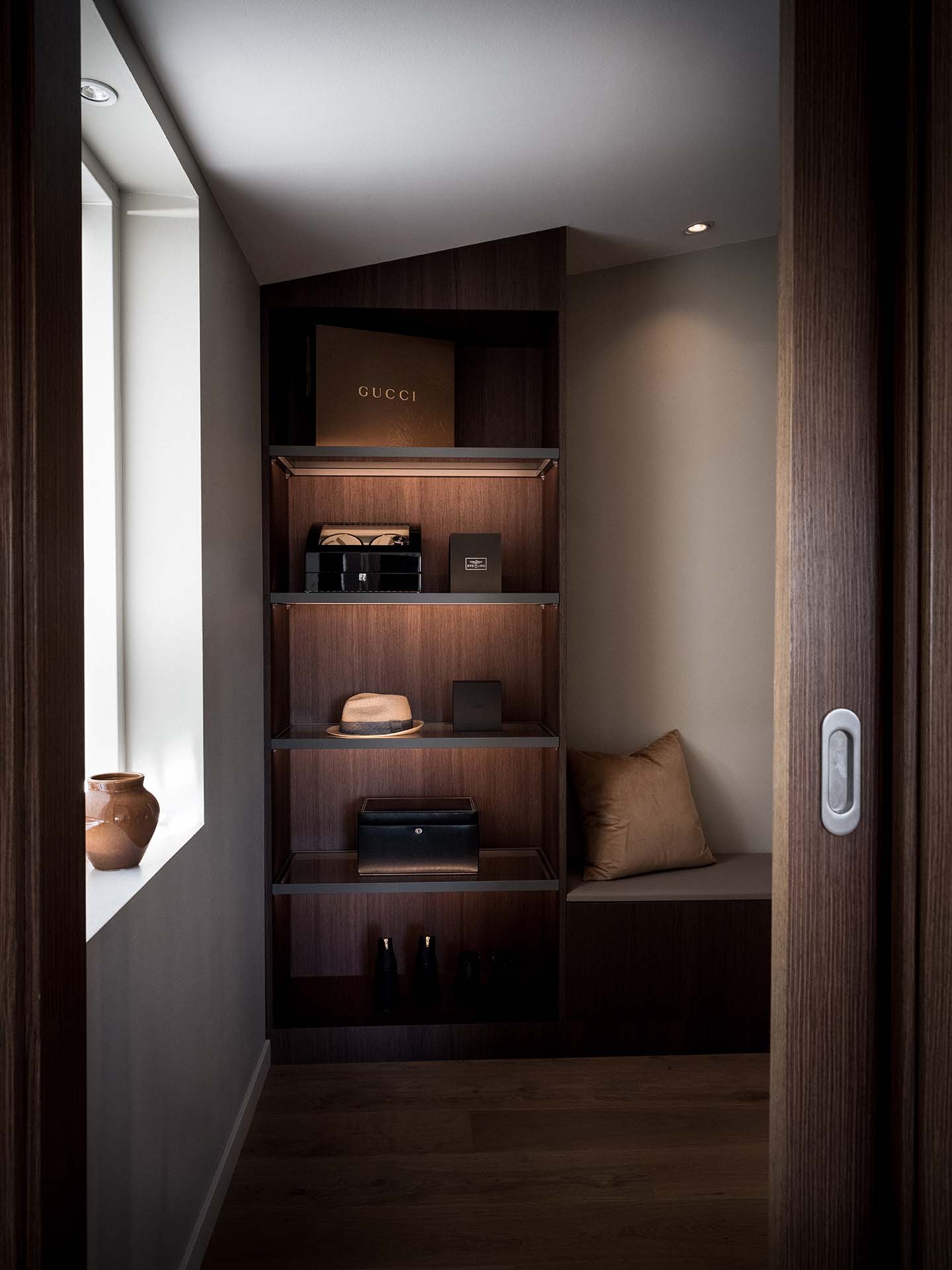 Stiligt - En liten modern alkov med inbyggda trähyllor som visar en hatt, väskor, lådor och inredning, bredvid en dämpad bänk och ett fönster med naturligt ljus.