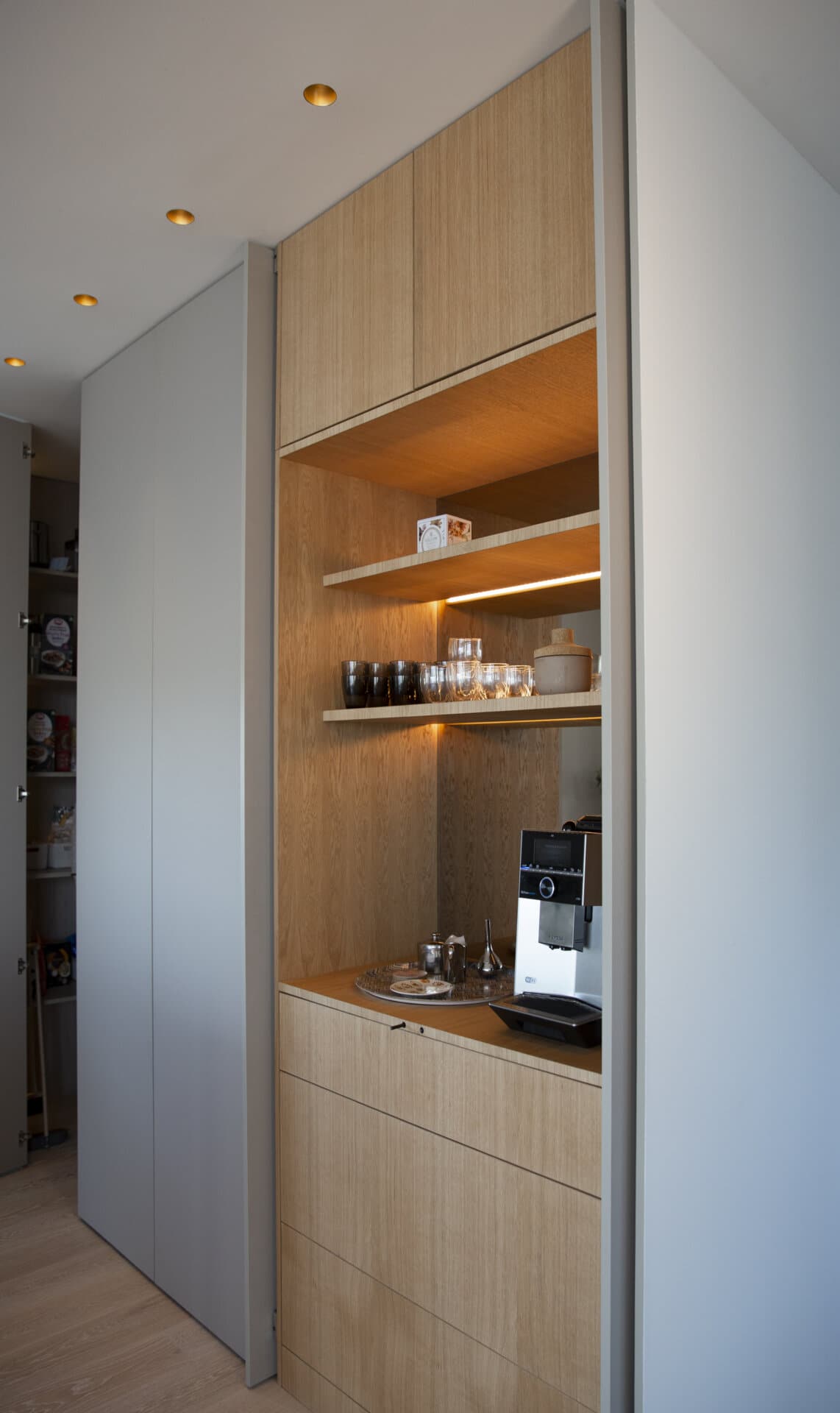 Stiligt - Fernströms minimalistiska kök En stiligt kökshörn har trähyllor med glasögon, en kaffemaskin och en bricka med koppar, allt elegant inställt mot en bakgrund av ljust trä och grå panel.