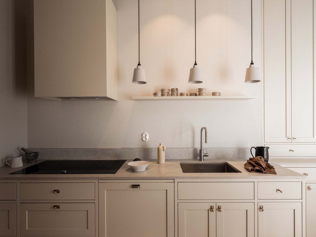Stiligt - Minimalistiskt kök med beige skåp, diskho, spishäll och taklampor. Öppen hylla ovanför rymmer flera koppar och skålar. Bänkskivan har en tekanna, skål och vattenkokare med handduk.