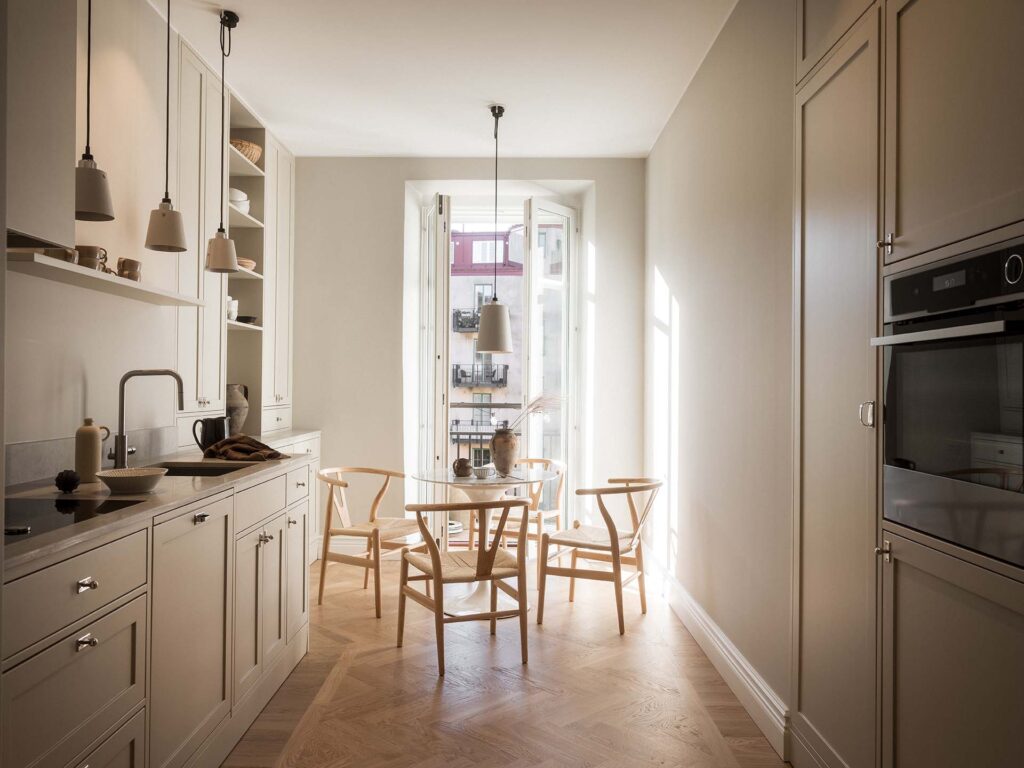 Stiligt - Minimalistiskt kök med ljusa träskåp, inbyggda vitvaror och ett runt matbord för fyra. Stora fönster släpper in naturligt ljus – perfekt när du väljer färg till ditt kök för en ljus och inbjudande atmosfär.