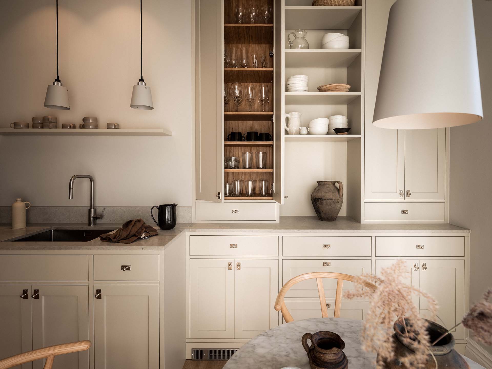 Stiligt - Minimalistiskt kök med krämskåp, öppna hyllor med porslin och glasföremål, ett handfat och en neutral inredning. Runt matbord med stolar och torkade blommor i förgrunden.