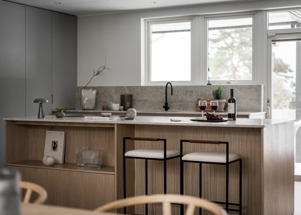 Stiligt - Modernt kök med ljusa träskåp, två barstolar, en bänkskiva i marmor och en flaska vin med två glas nära ett handfat under stora fönster.