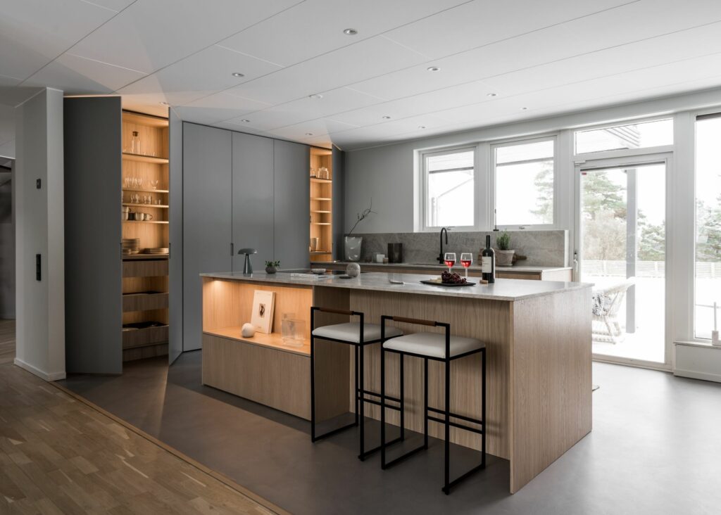 Stiligt - Modernt kök med centralö, två barstolar, inbyggda hyllor och stora fönster som släpper in dagsljus. Neutral färgpalett med trä och grå finish – perfekt inspiration när du väljer färg till ditt kök.