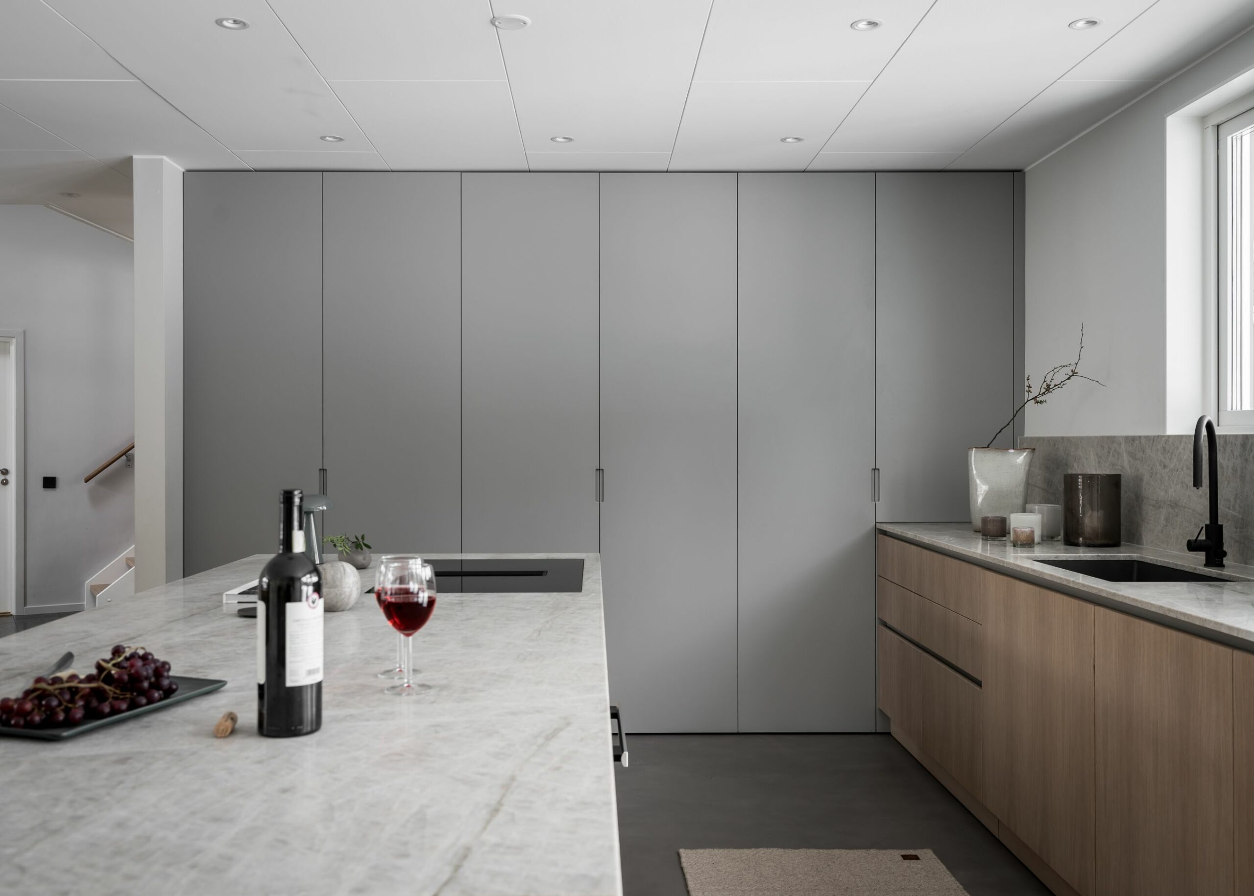 Stiligt - Modernt kök med minimalistiska gråa skåp, bänkskivor i marmor, en köksö med vin och vindruvor och ett fönster som släpper in naturligt ljus.
