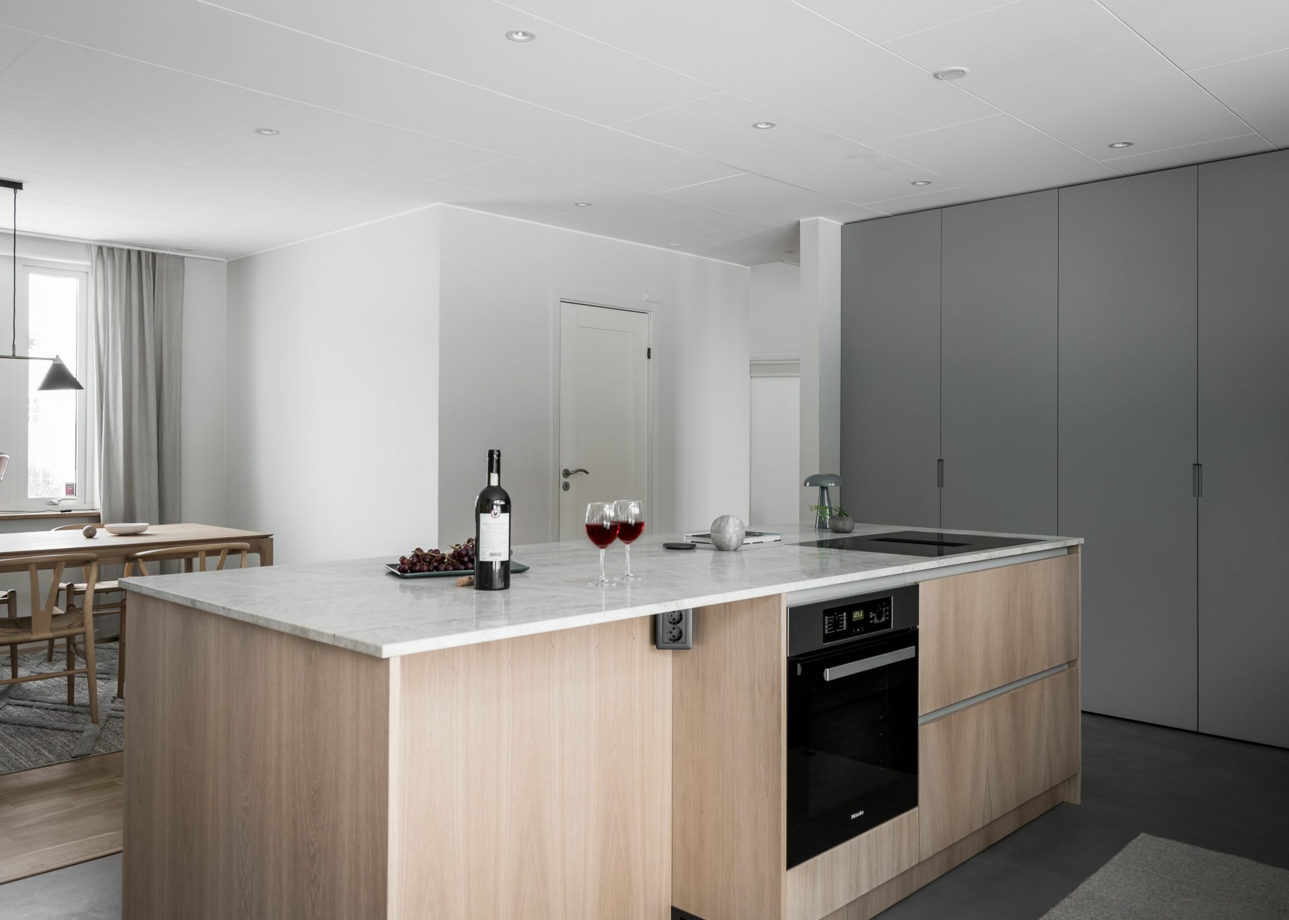 Stiligt - Modernt kök med bänkskiva i marmorö, inbyggd ugn, två glas rött vin, en flaska och vindruvor. Minimalistisk matplats med träbord synligt i bakgrunden.