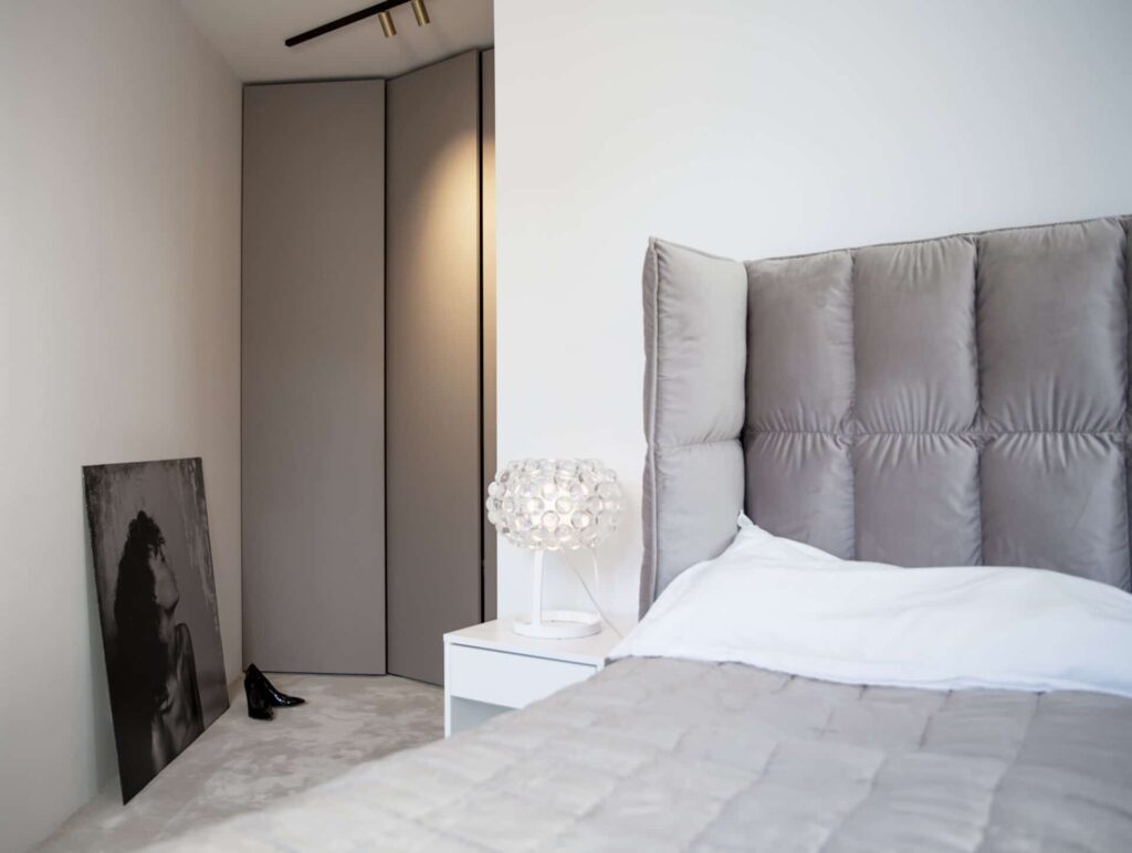 Stiligt - Ett modernt sovrum med en tuftad grå sänggavel, vita sängkläder och ett nattduksbord med en sfärisk lampa visar upp Villa Fernströms sovrumsförvaring bredvid ett svartvitt porträtt på golvet nära hörngarderoben.