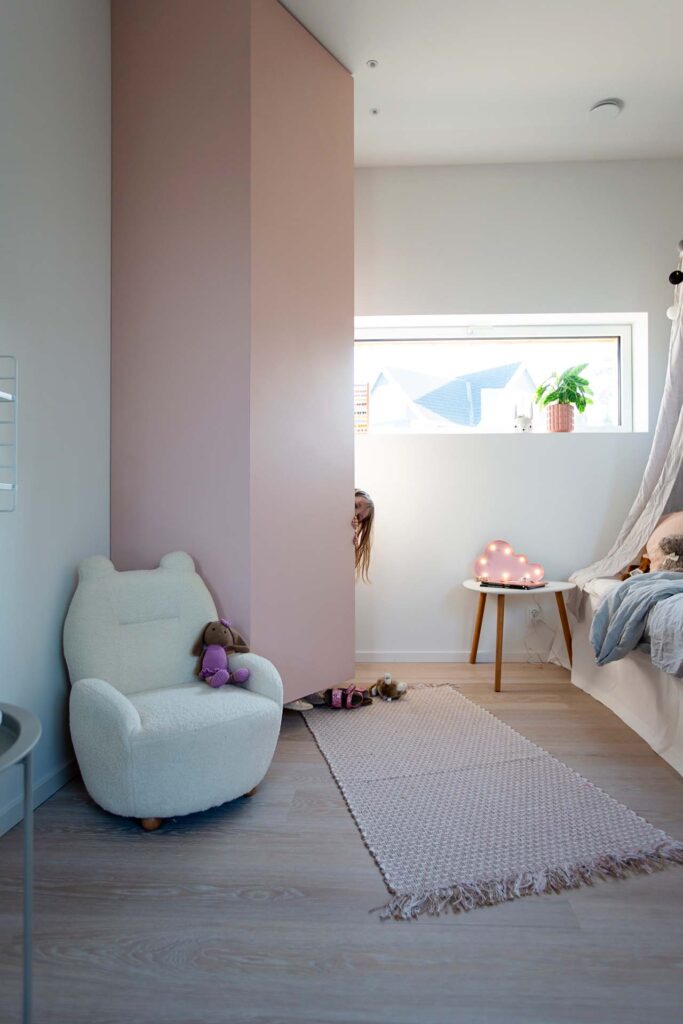 Stiligt - Villa Fernströms sovrumsförvaring. Ett barn kikar bakom en rosa vägg i ett stilrent, mysigt rum med en plyschstol, liten matta, sidobord och säng.