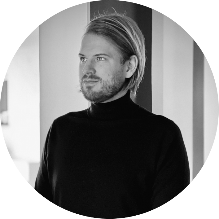 Stiligt - En man med ljust hår och skägg, klädd i svart polokrage, står inomhus och tittar åt sidan. Bilden är i svartvitt.