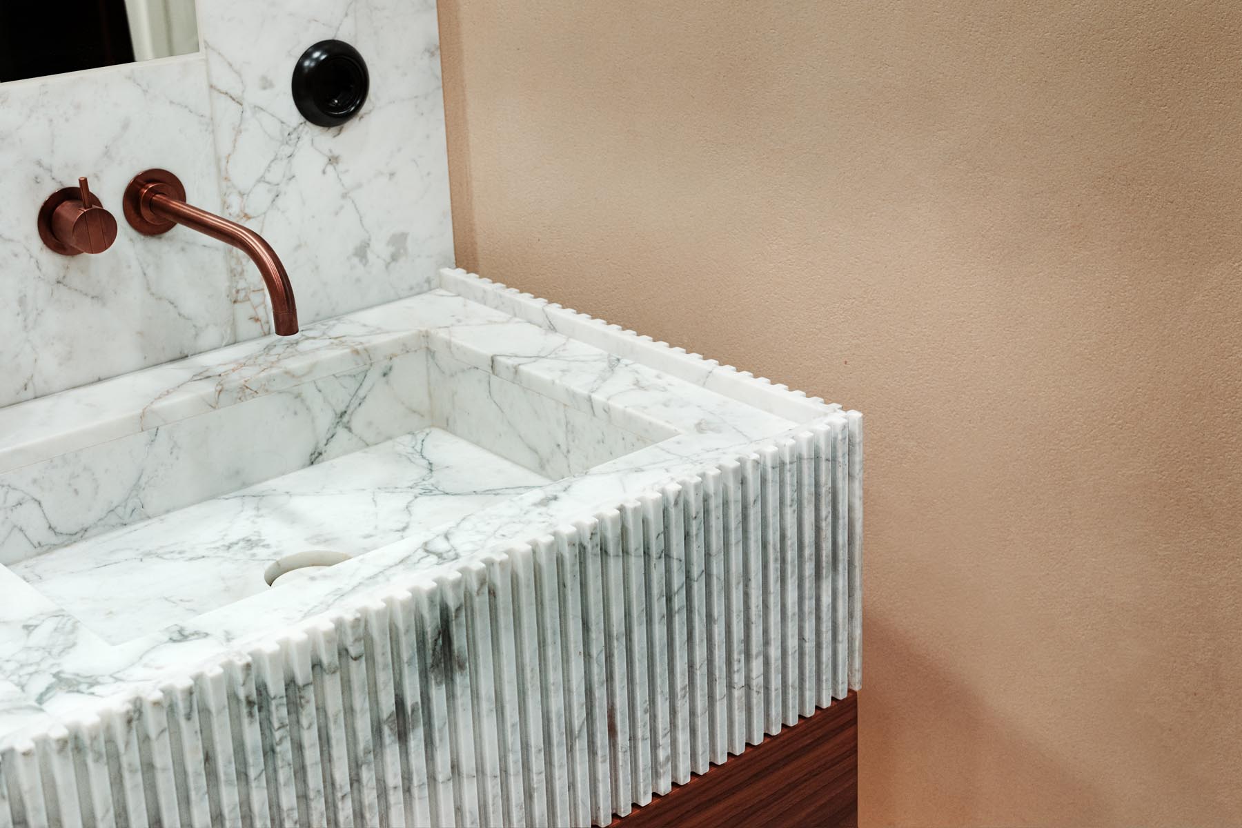 Stiligt - Ett handfat i marmor med en räfflad frontdesign, en väggmonterad kopparblandare och en beige vägg skapar en lyxig badrumsatmosfär.