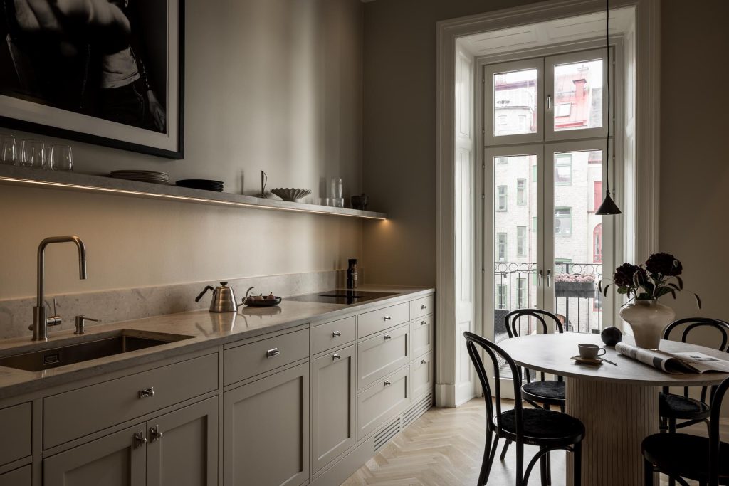 Stiligt - Modernt kök i kök i shakerstil med ljusa skåp, bänkskiva i marmor, belysning under hyllplan, diskho och ett matbord med stolar bredvid stora fönster med utsikt över en stadsgata.