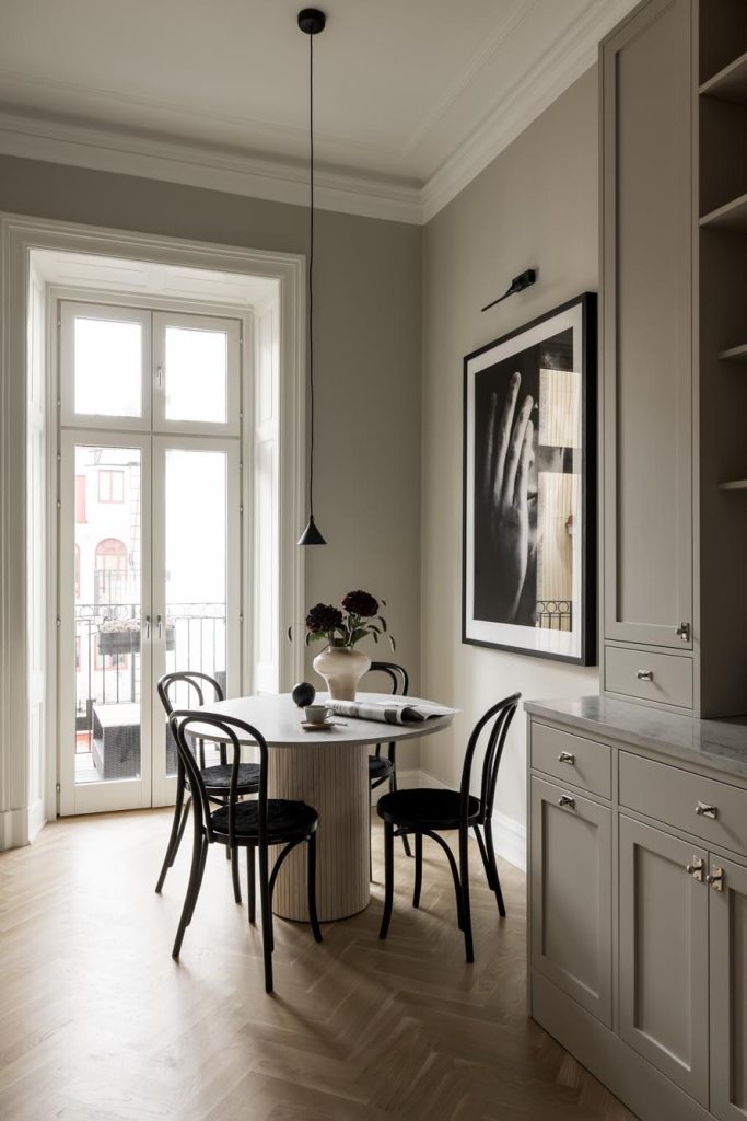 Stiligt - En modern matplats med ett runt bord, fyra svarta stolar, en blommig mittpunkt och ett slående svart-vitt handfotografi på väggen, allt nära höga fönster och inbyggnadsskåp i kök i shakerstil.