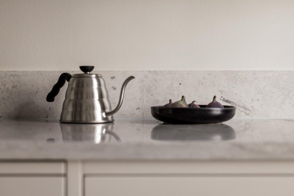 Stiligt - En svanhalsvattenkokare i rostfritt stål och en svart skål med fikon sitter på en bänkskiva i marmor i en kök i shakerstil, ställd mot en ljus vägg.