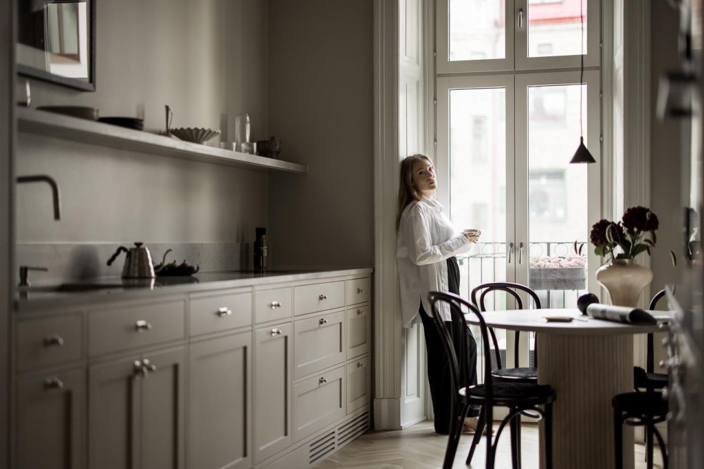 Stiligt - En kvinna står vid ett stort fönster i ett modernt kök i shakerstil, håller i en kopp och tittar ut. Köket har neutrala toner, ett matbord och blommor i en vas.