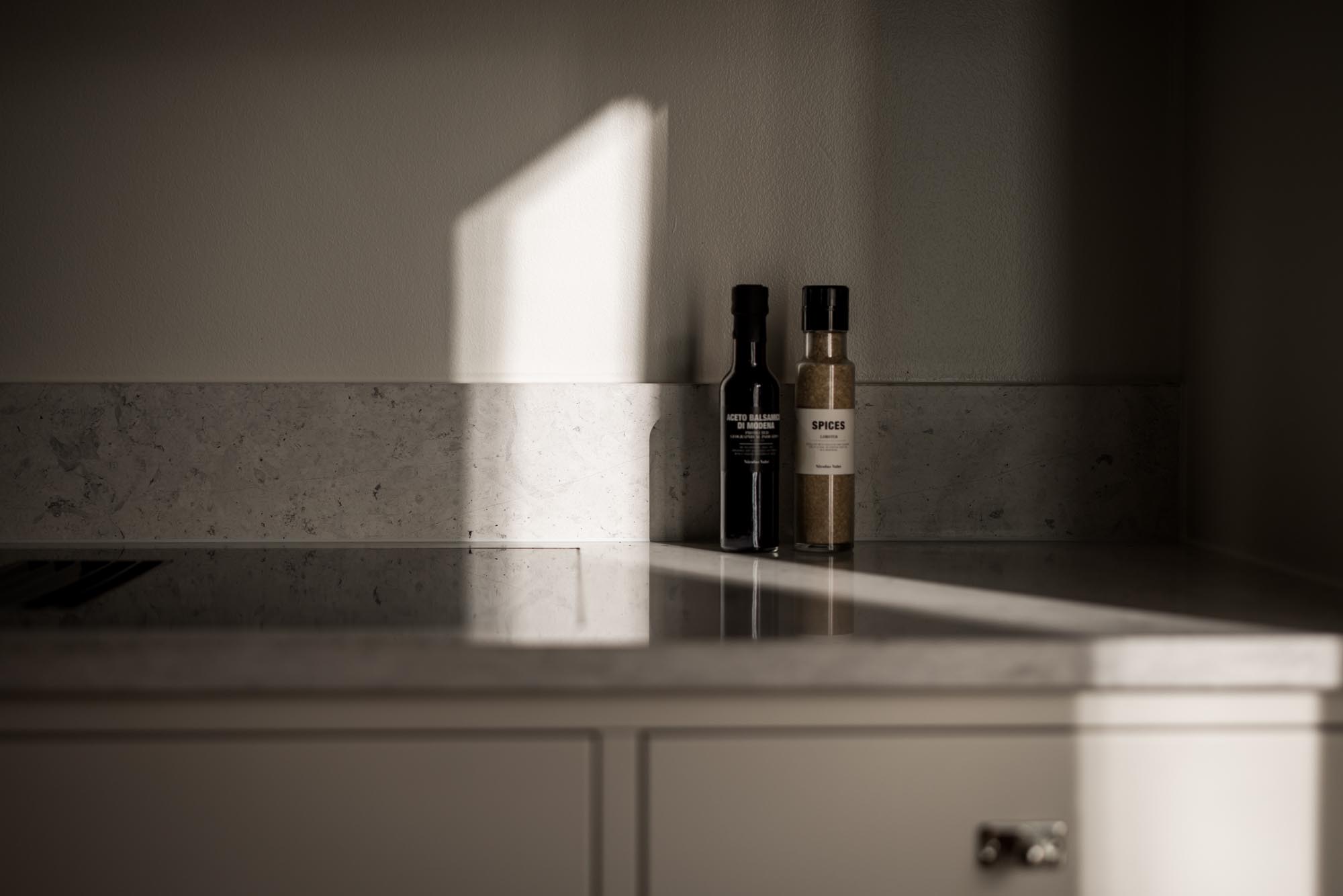 Stiligt - Två flaskor, en mörk och en ljus, sitter på en bänkskiva i marmor i en kök i shakerstil, när solljuset strömmar in och kastar mjuka skuggor på vägg och yta.