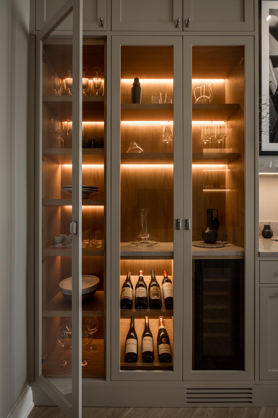 Stiligt - Skåp med glasfront i kök i shakerstil med interiörbelysning visar vinflaskor, glas, karaffer och prydnadsföremål, tillsammans med en inbyggd vinkyl.