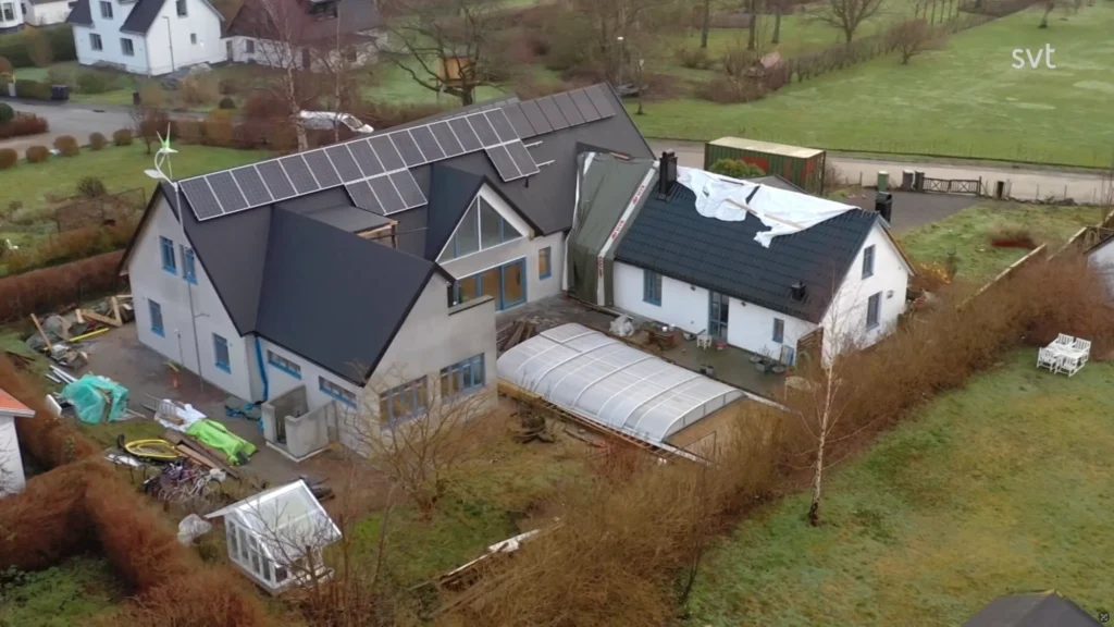 Stiligt - Flygfoto över två sammankopplade hus, ett med solpaneler och det andra med ett delvis skadat tak; Det omgivande området inkluderar träd, ett växthus och trädgårdsutrustning.