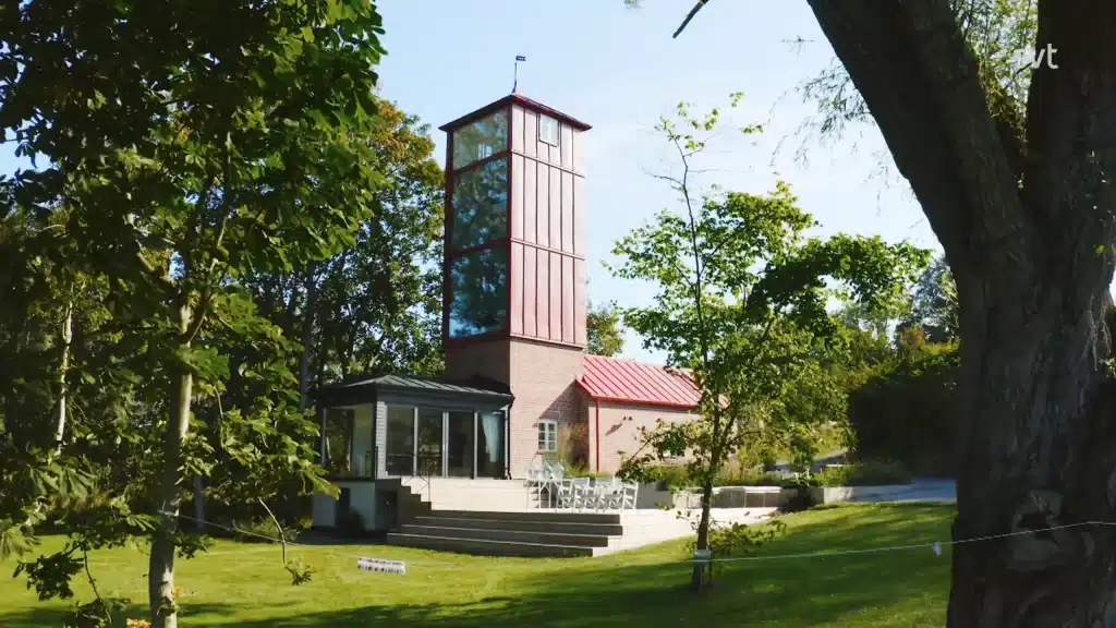 Stiligt - Ett modernt hus med ett högt glastorn och rött tak står bland gröna träd och en gräsbevuxen gräsmatta en solig dag.