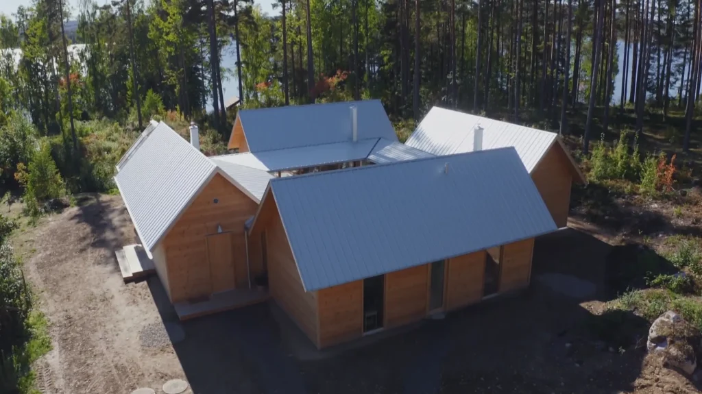 Stiligt - Flygfoto över ett kluster av fyra trähytter med plåttak omgivna av träd, med en glimt av vatten i bakgrunden.