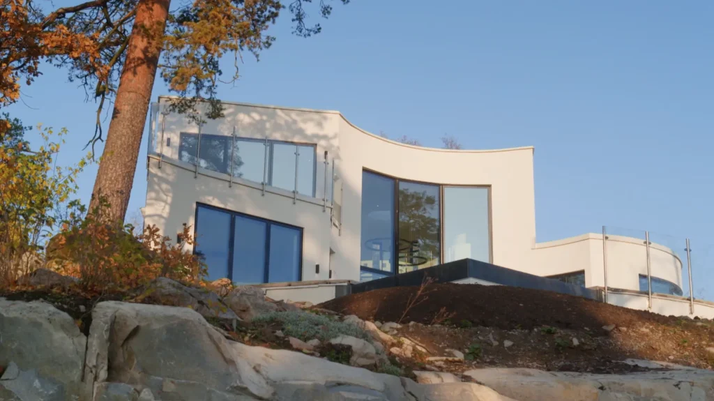 Stiligt - Modernt vitt hus med stora glasfönster och en svängd design, beläget på en stenig sluttning under en klarblå himmel.