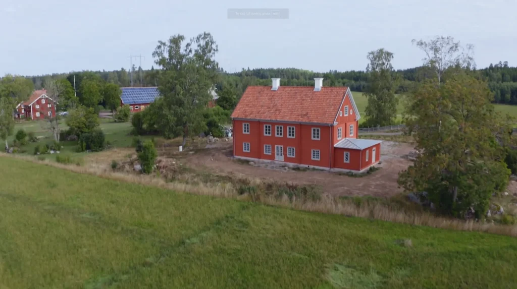 Stiligt - Ett rött tvåvåningshus med vita detaljer står i ett lantligt område omgivet av åkrar och träd, med ytterligare byggnader synliga i bakgrunden.
