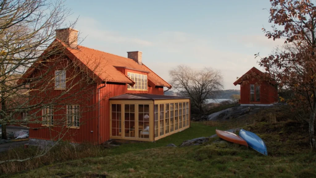 Stiligt - Ett rött trähus med solrumsförlängning i glas står på en gräsbevuxen gård nära en mindre röd byggnad, med kajaker och träd i närheten.
