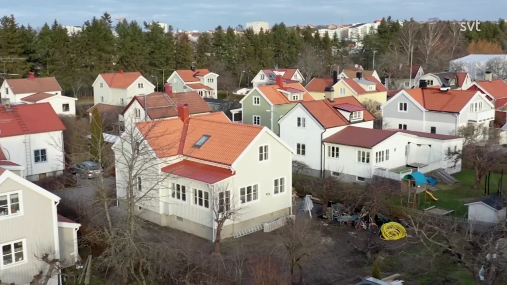 Stiligt - Flygfoto över ett bostadsområde med vita hus med röda tak, omgivna av träd, under en klar himmel.