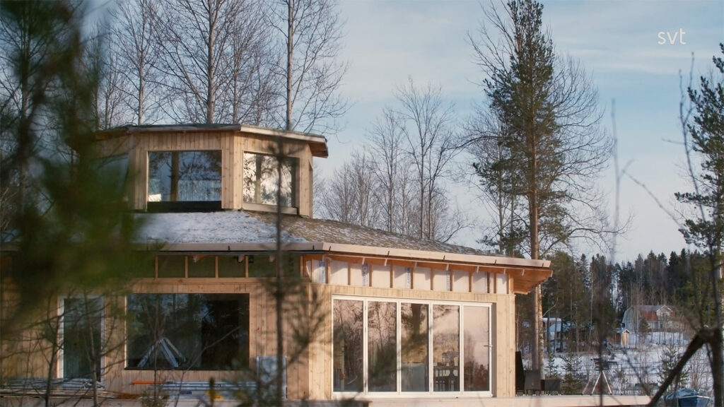 Stiligt - Ett trähus med stora fönster och en liten övre struktur, omgivet av bladlösa träd i ett snölandskap under en klar himmel.
