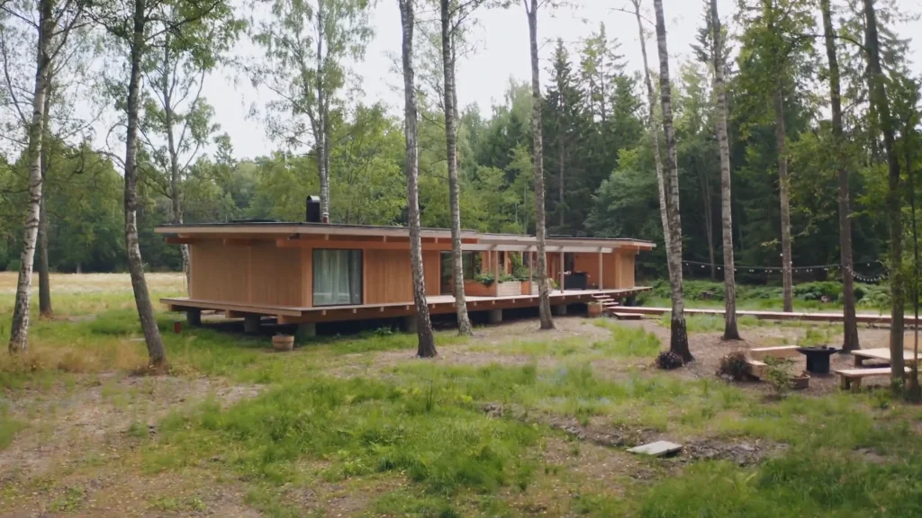 Stiligt - Ett modernt trähus med stora fönster ligger bland höga träd i en skogsglänta, med ett rymligt däck och omgivet av grönska.