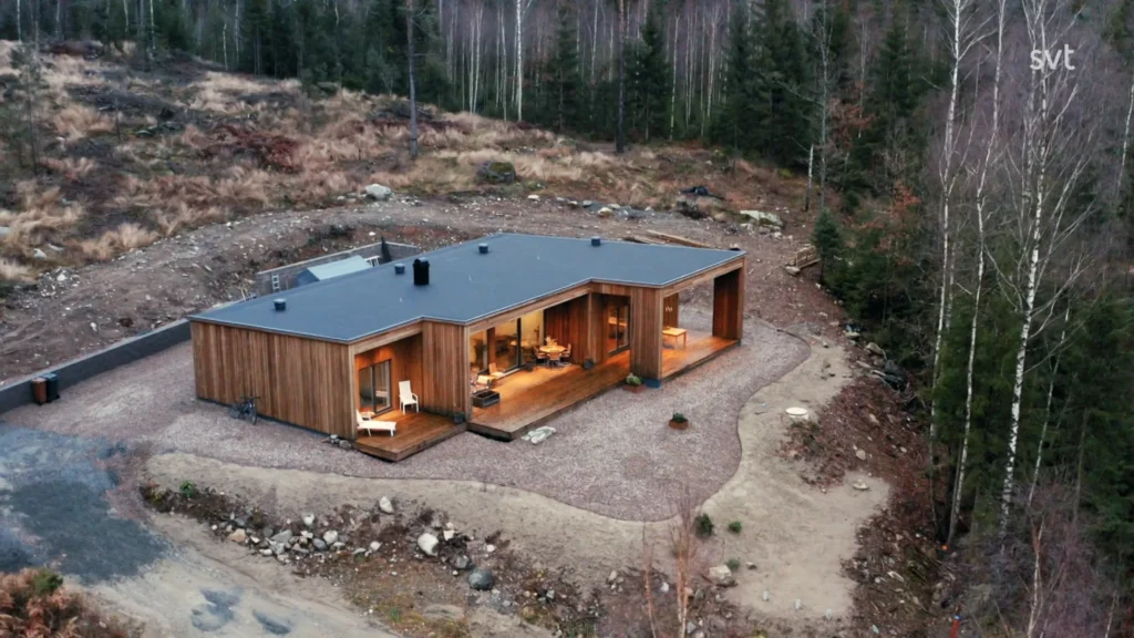 Stiligt - Ett modernt enplans trähus med stora fönster ligger i en skogsglänta med omgivande träd och stenig terräng.