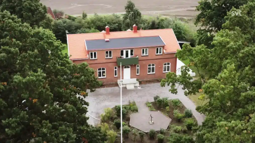 Stiligt - Flygfoto över ett tvåvåningshus i rött tegel med tegeltak, omgivet av träd och en anlagd trädgård med ett cirkulärt inslag framför.