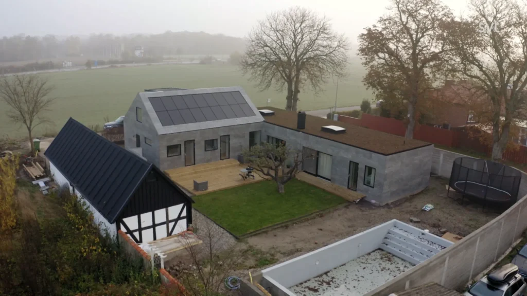 Stiligt - Flygfoto över ett modernt hus med solpaneler på taket, ett fristående svartvitt uthus, en gräsmatta på bakgården och en tom pool. Träd och dimmiga fält omger fastigheten.