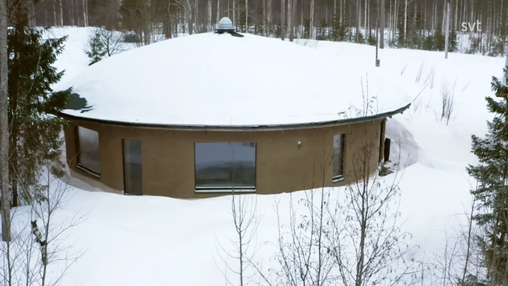 Stiligt - Ett runt, jordfärgat hus med stora fönster är täckt av snö och omgivet av ett snöigt skogslandskap.