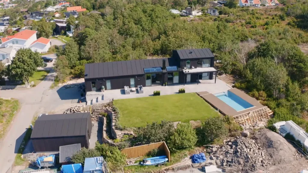 Stiligt - Flygfoto över ett modernt svart hus med en stor gräsmatta, pool och omgivande grönska i ett bostadsområde.