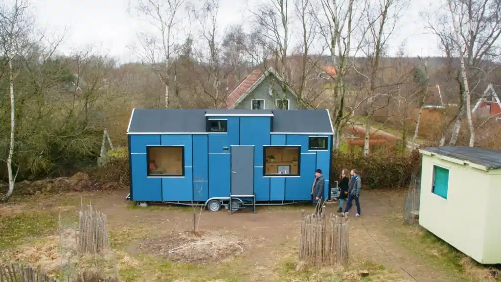 Stiligt - Ett modernt blått litet hus på hjul är parkerat på en gräsbevuxen tomt, med tre personer som står i närheten. Bladlösa träd och hus syns i bakgrunden.