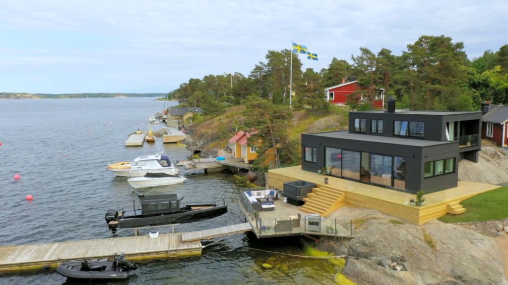 Stiligt - Modernt svart hus med stora fönster med utsikt över en brygga vid vattnet med båtar; Svensk flagga synlig bland träd och ytterligare hus i närheten.